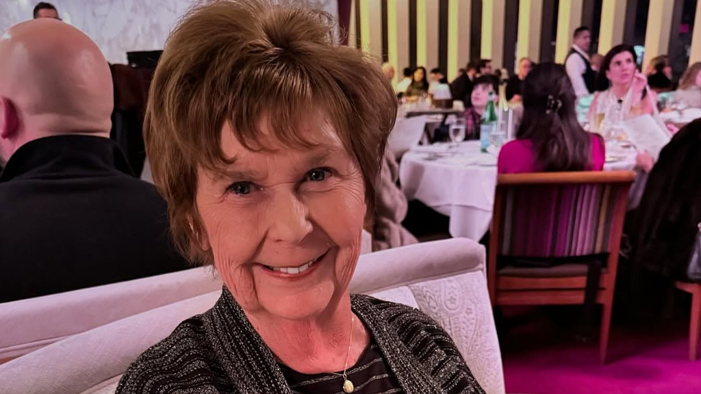 A smiling older woman with brown hair and bangs looks directly at the camera, wearing a sparkly dark cardigan and a pendant necklace, sitting on a light upholstered bench. Behind her, in a blurred restaurant setting, other diners sit at white-clothed tables, a waiter stands, and a wall with vertical dark and light stripes is visible. The carpet is a bright fuchsia.