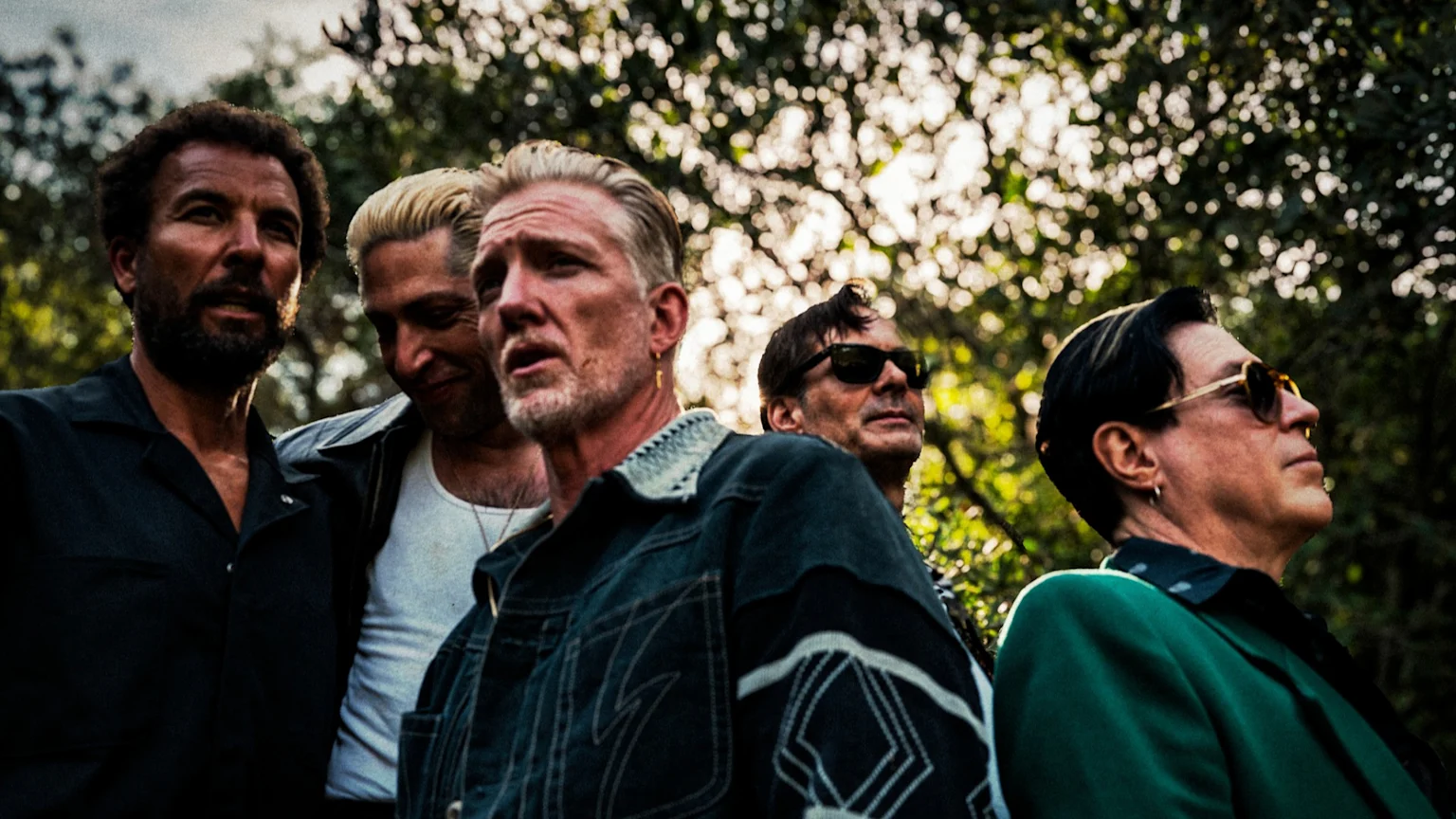 Five men stand outdoors, partially backlit by sunlight filtering through tree leaves, with some looking forward and others looking up or to the side.