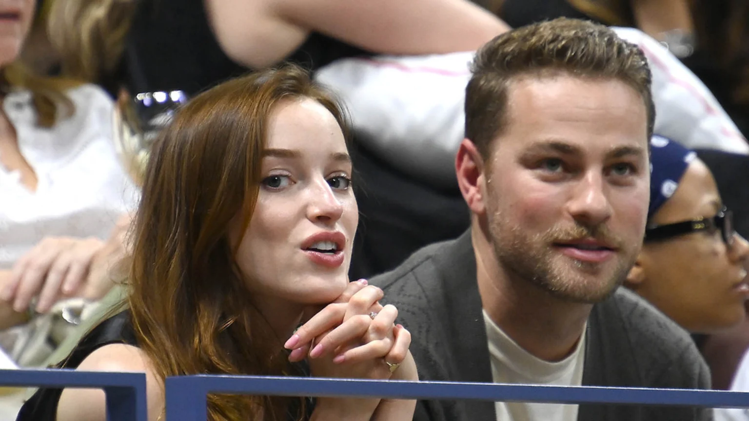 Phoebe Dynevor, showing a ring on her left ring finger, sits beside Cameron Fuller in a public setting.
