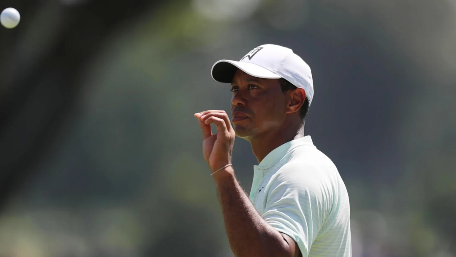 Golfer Tiger Woods, wearing a white cap and light green polo shirt, looks intently to his left, with a blurred white golf ball visible in the upper left.