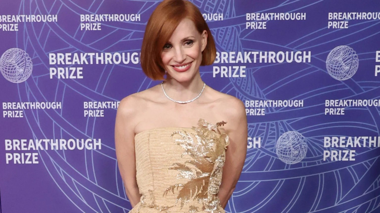 Jessica Chastain, with red hair and a diamond necklace, smiles in a strapless gold dress against a purple "BREAKTHROUGH PRIZE" backdrop.