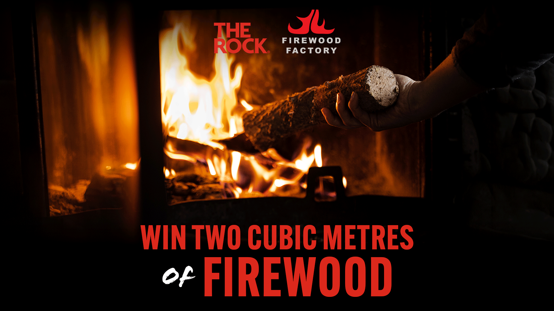 A hand places a log into a blazing wood stove with bright orange flames, above text reading "WIN TWO CUBIC METRES of FIREWOOD" and "THE ROCK." and "FIREWOOD FACTORY" logos.