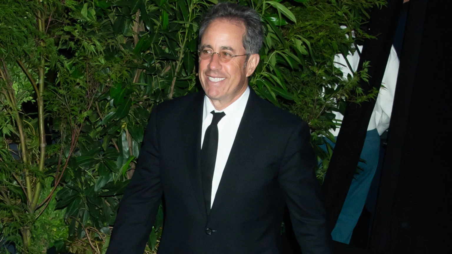 Jerry Seinfeld smiles at the camera, wearing a black suit, white shirt, and black tie, with green foliage in the background.