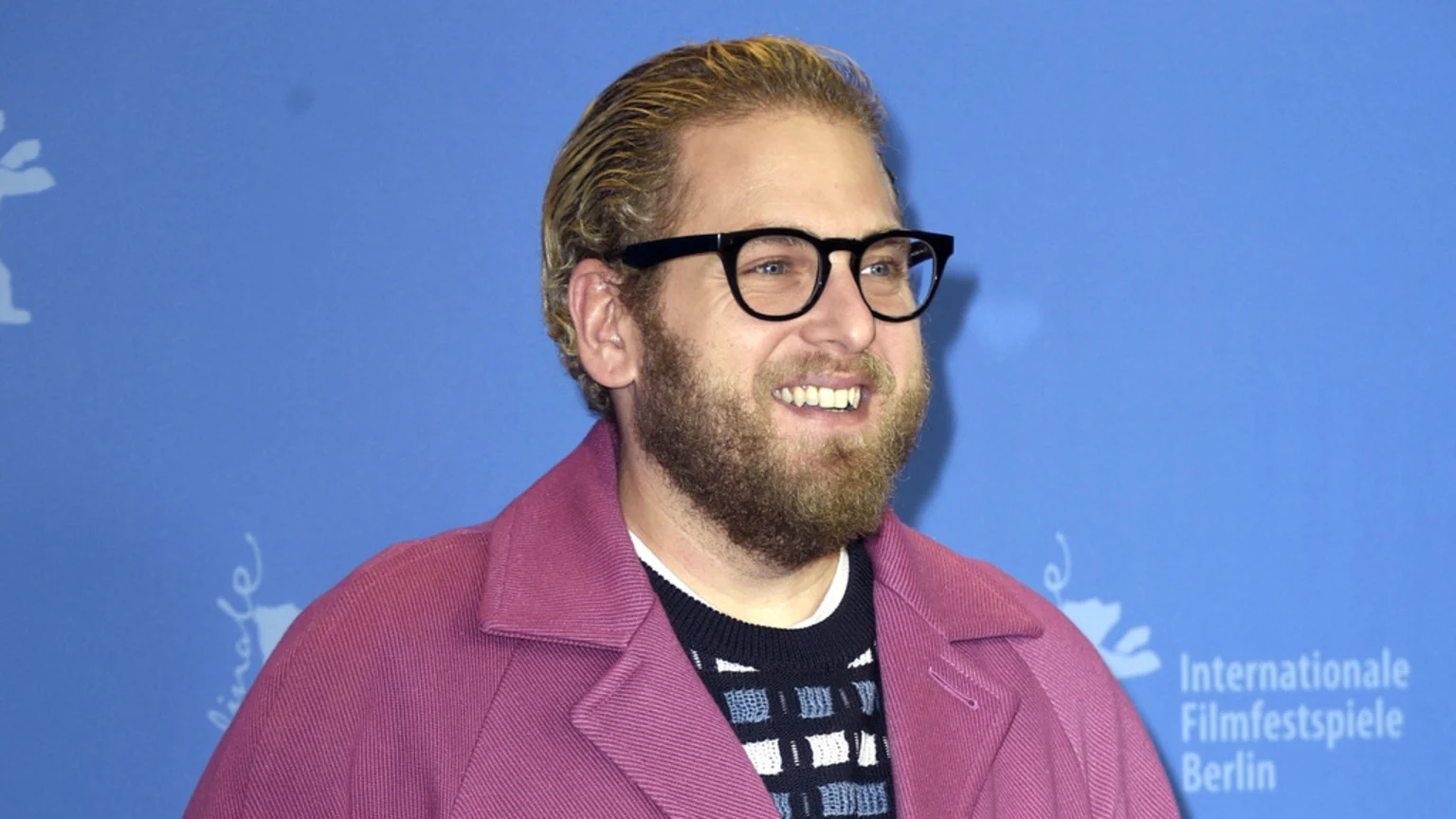 Jonah Hill smiles, wearing black glasses, a blonde beard, and a pink coat over a patterned sweater, against a blue background with the Berlin Film Festival logo.