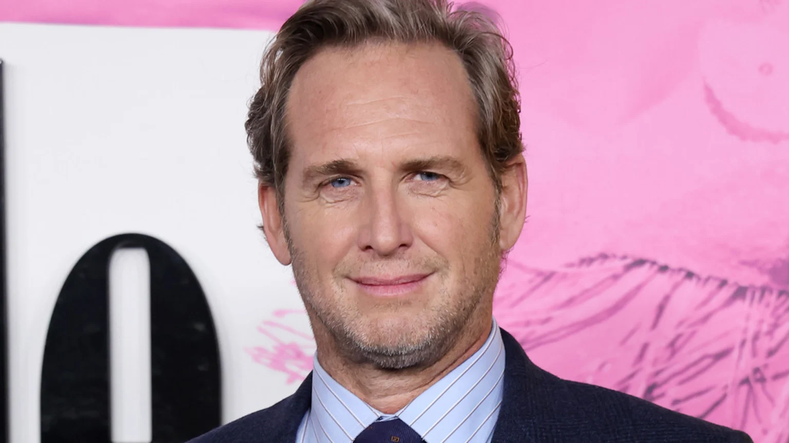 A smiling man with light brown hair, blue eyes, and stubble wears a blue pinstripe shirt, dark tie, and jacket, against a white background with a black "0" and a pink patterned section.