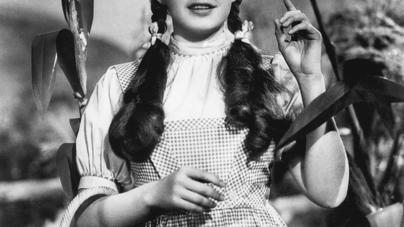 A black and white photo of a young Judy Garland as Dorothy, wearing a gingham dress and pigtails, holding a basket, and pointing upwards amidst cornstalks.