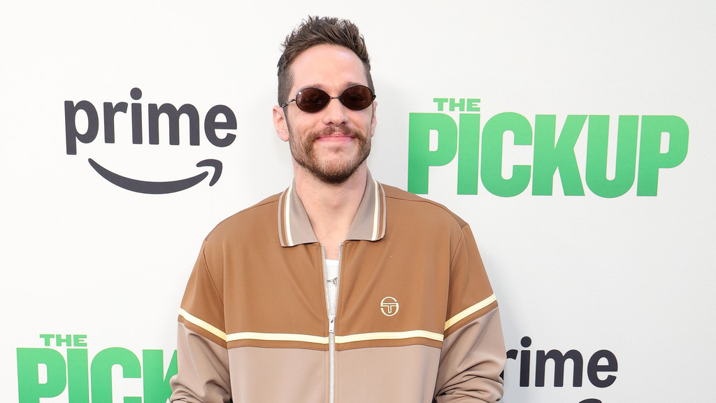 Pete Davidson smiles slightly, wearing small sunglasses and a brown track jacket with light stripes, standing in front of a white background with "prime" and "THE PICKUP" logos.