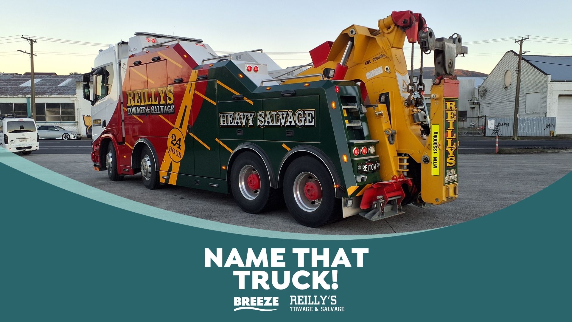 A large, multi-colored Reilly's Towage & Salvage heavy wrecker, painted white, red, green, and yellow, is parked on asphalt with industrial buildings in the background.