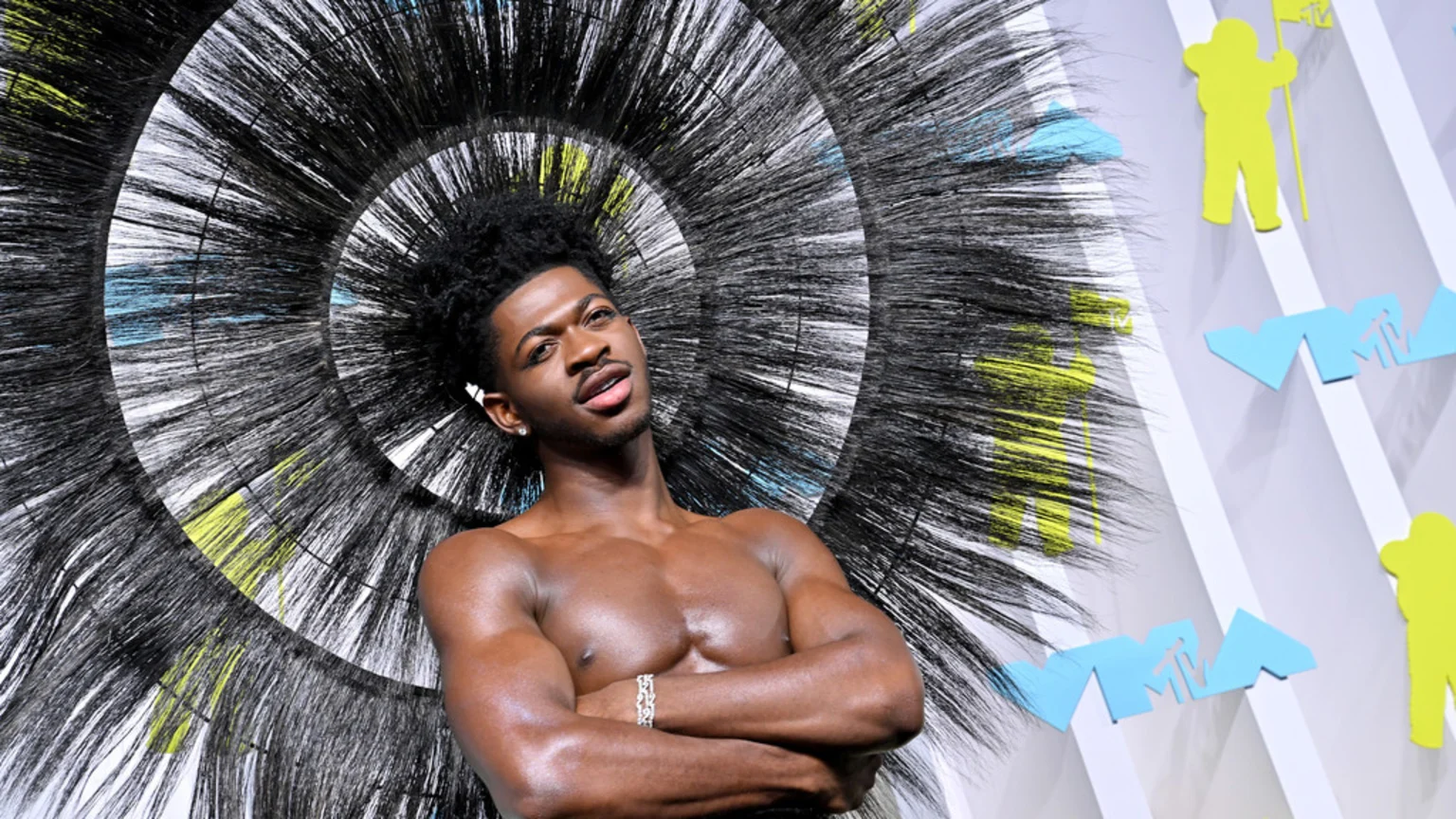 Lil Nas X, shirtless with crossed arms, poses against a striking black spiky circular backdrop with hints of yellow and blue, and light blue and yellow MTV logos on a white wall.