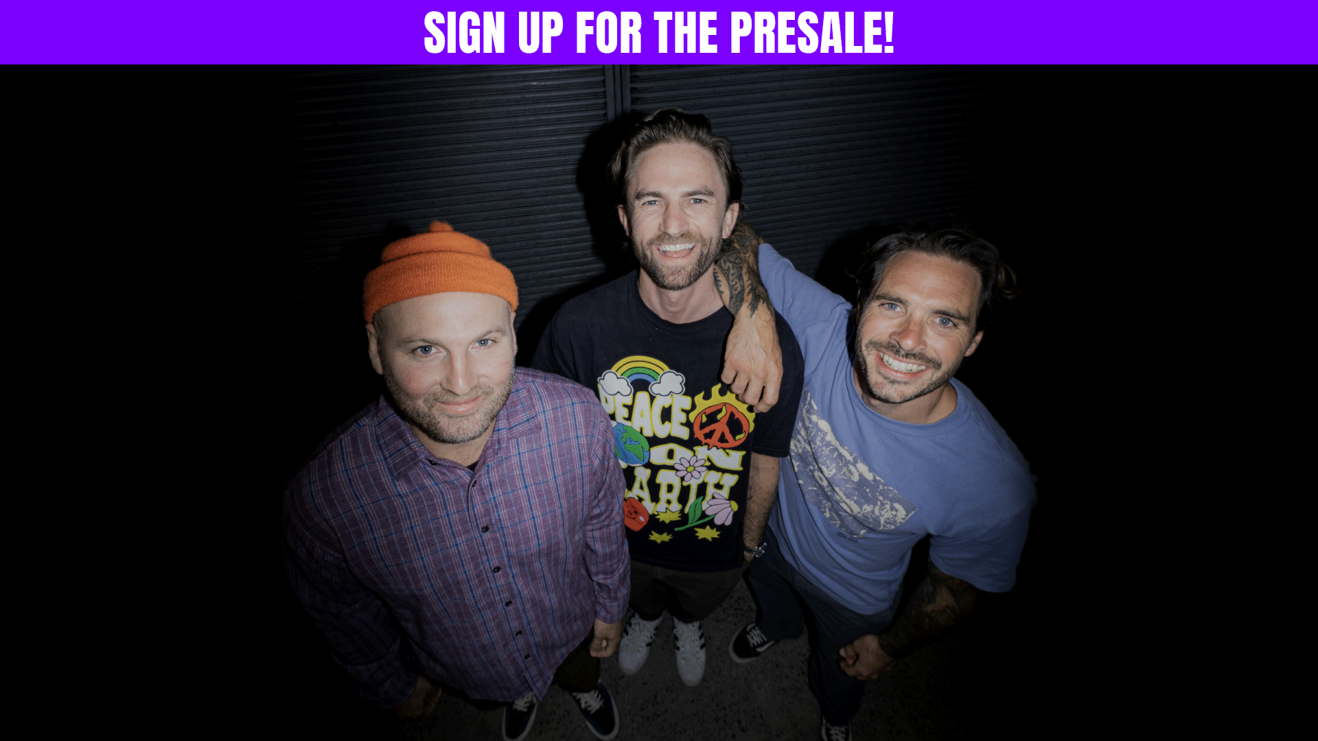 Three smiling men look up at the camera; one in an orange beanie and plaid shirt, the middle man in a "PEACE ON EARTH" shirt with his arm around the third man in a blue shirt with tattoos.