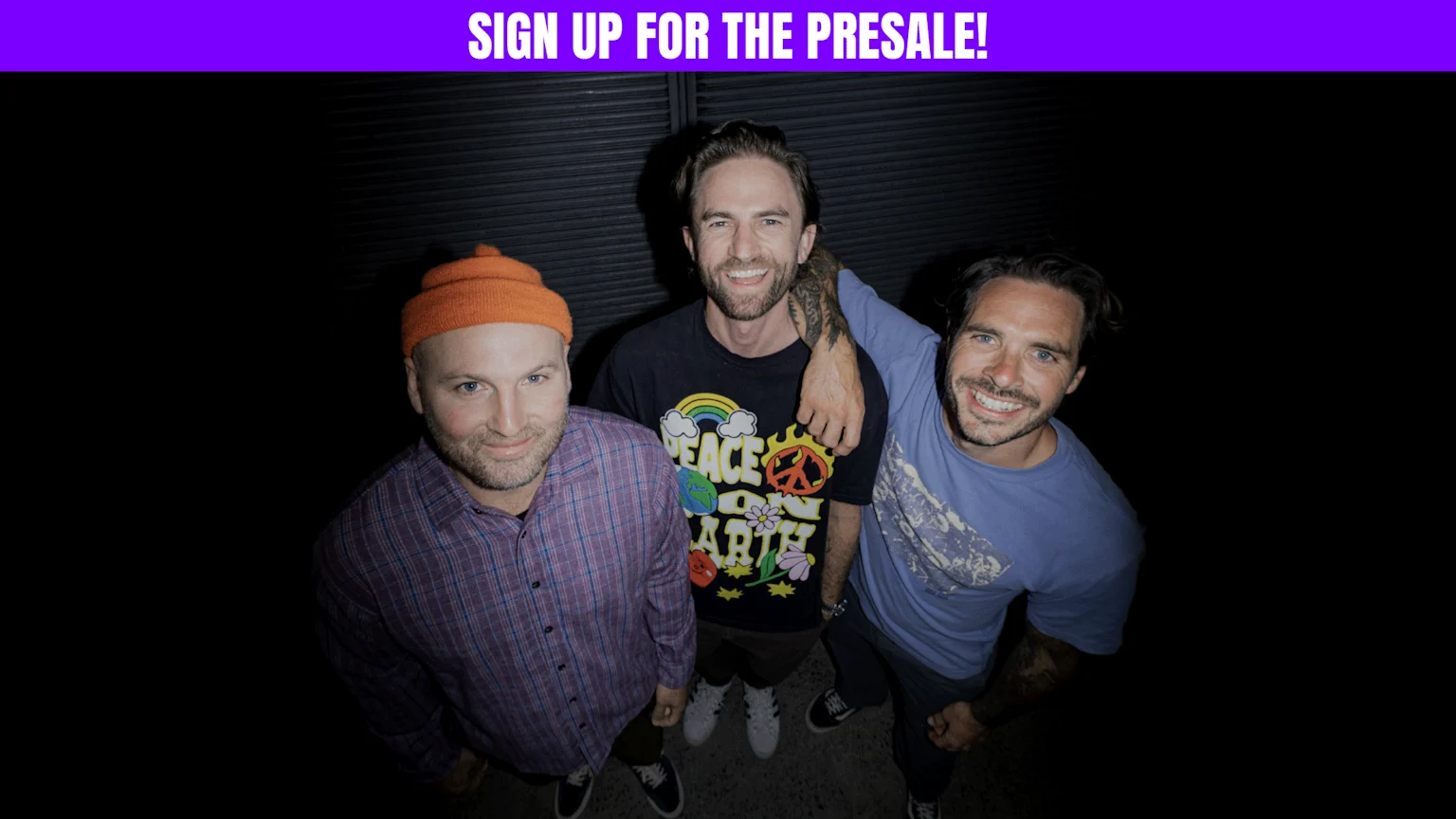 Three smiling men look up at the camera; one in an orange beanie and plaid shirt, the middle man in a "PEACE ON EARTH" shirt with his arm around the third man in a blue shirt with tattoos.