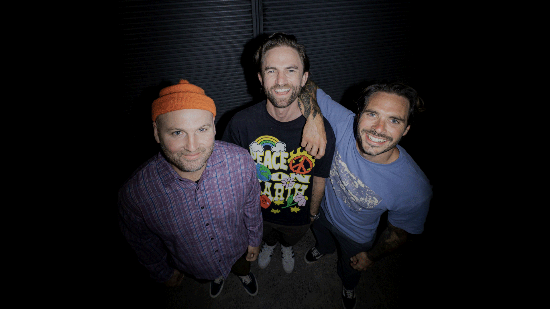 Three smiling men look up at the camera; one in an orange beanie and plaid shirt, the middle man in a "PEACE ON EARTH" shirt with his arm around the third man in a blue shirt with tattoos.