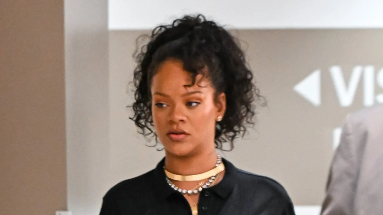 Close-up of Rihanna, wearing a black collared shirt and layered gold and diamond necklaces, looking to the side with her curly hair pulled back.
