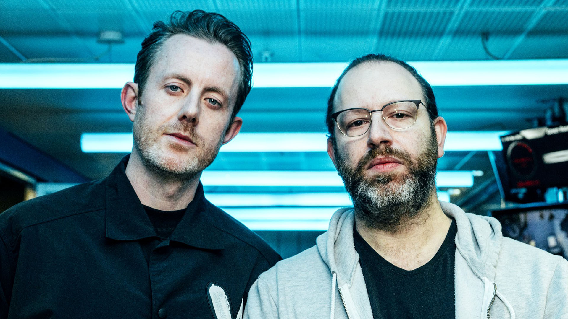 Two serious-looking men, one with short dark hair and a beard in a dark shirt, and the other with glasses and a full beard in a gray hoodie, look at the camera against a blue-lit background.
