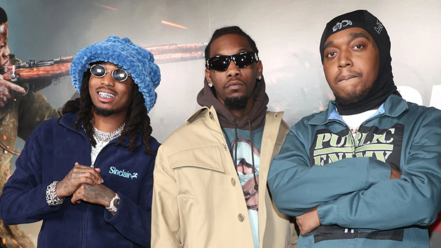 Three members of Migos stand together: Quavo on the left in a blue fuzzy hat and navy jacket, Offset in the middle in sunglasses and a tan jacket, and Takeoff on the right in a black balaclava and teal jacket with "Public Enemy" logo.