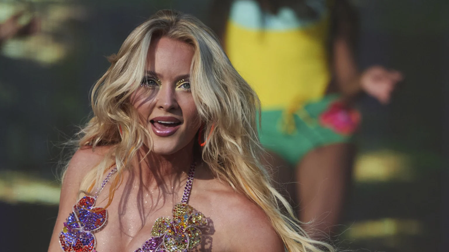A blonde woman with long, wavy hair, glittery yellow eye makeup, and a colorful jewel-encrusted top smiles at the camera, with a blurred figure in a yellow and green outfit behind her.