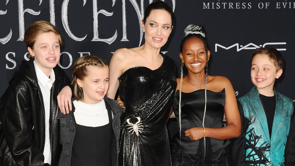 Angelina Jolie, in a black dress with a scorpion brooch, poses with her smiling daughter Zahara and three other children on a black background.