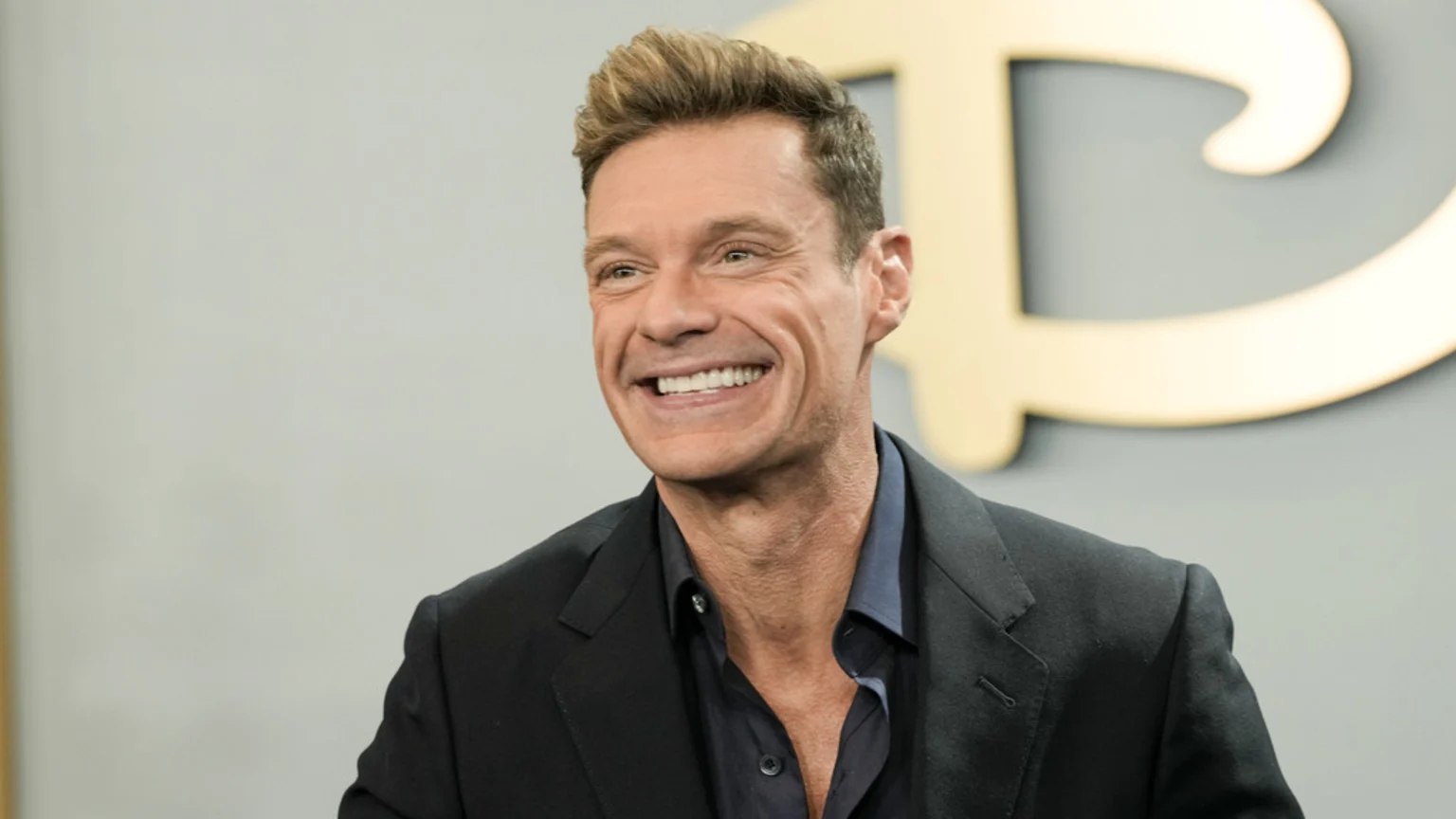A smiling Ryan Seacrest, a man with light brown hair, wears a dark suit jacket and blue shirt against a gray wall with a blurred golden letter in the background.