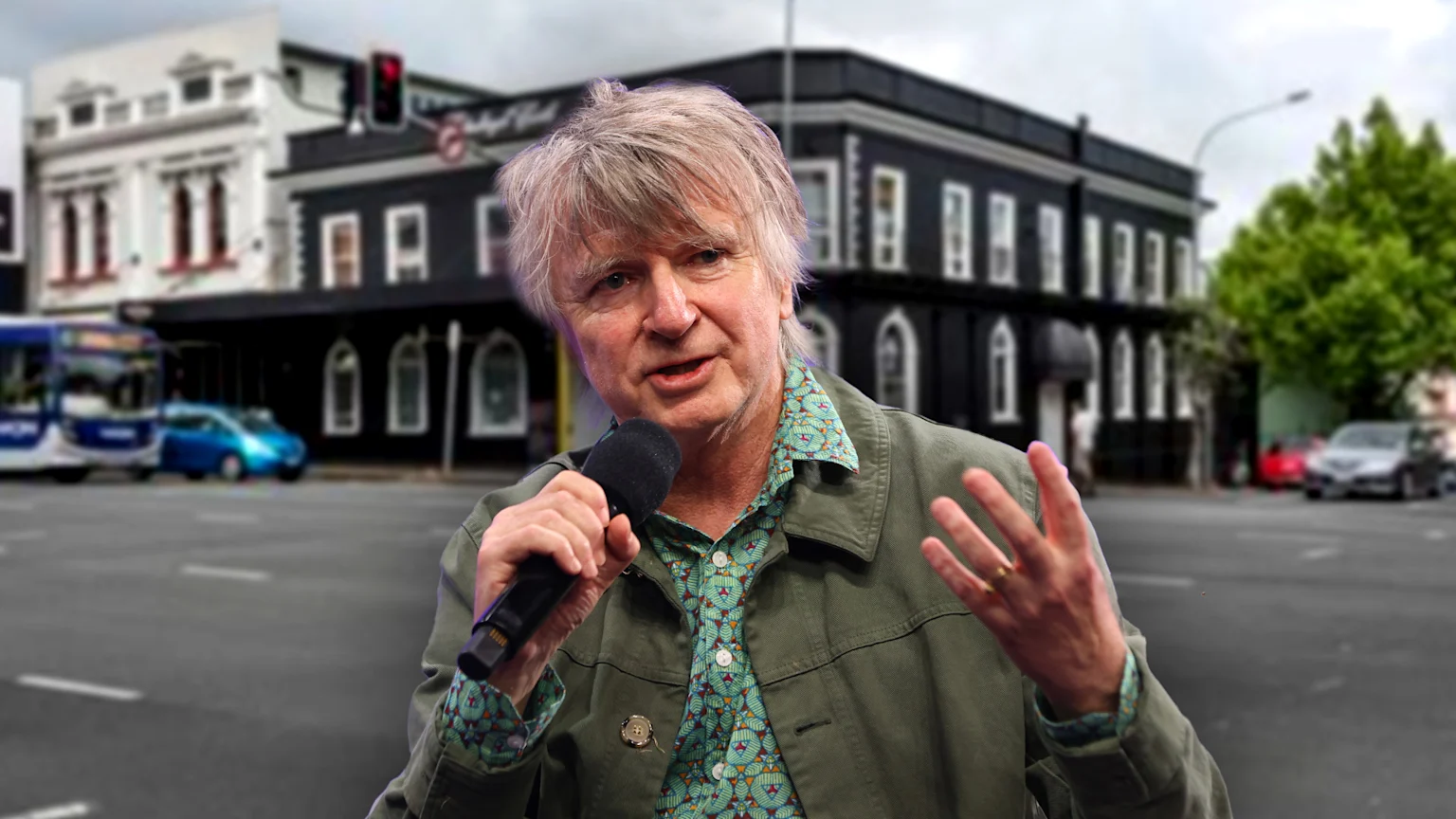 Musician Neil Finn holds a microphone and gestures, standing on a city street with blurred buildings, including a prominent dark one, and vehicles in the background.