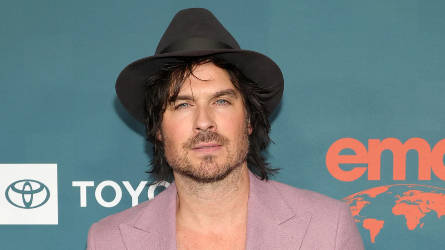 Ian Somerhalder with shaggy dark hair and a beard, wearing a dark hat and a pink blazer, stands against a teal background with white Toyota and orange EMC logos.