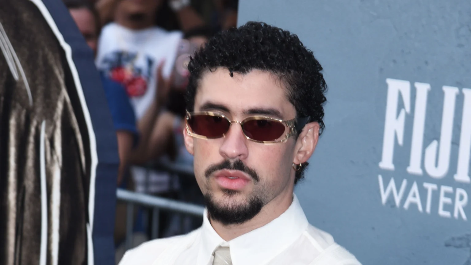 Bad Bunny wears gold-framed sunglasses with reddish-brown lenses, a light collared shirt and tie, a goatee, and a gold hoop earring, with a blurry "FIJI WATER" logo behind him.