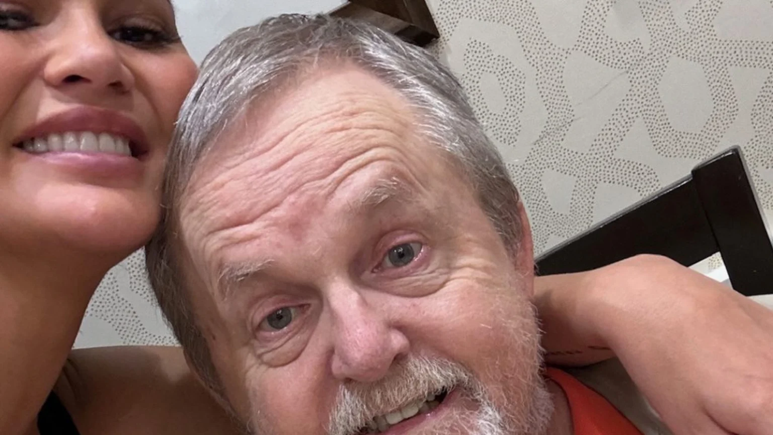 A close-up selfie shows a smiling woman with her arm around an older, smiling man with grey hair and a beard, against a patterned wallpaper background.