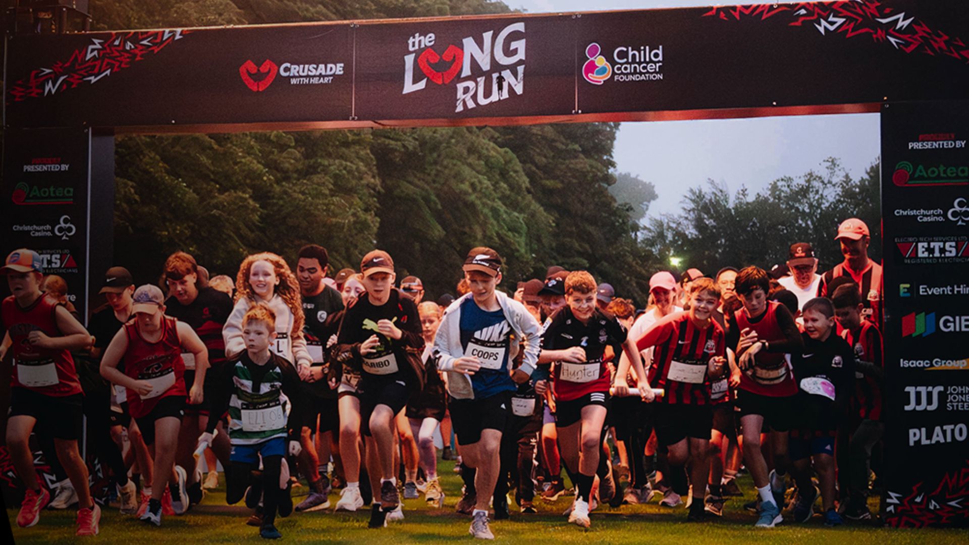 Children and adults begin a race on a grassy field, running under a large banner displaying "the LONG RUN" event title and sponsor logos.