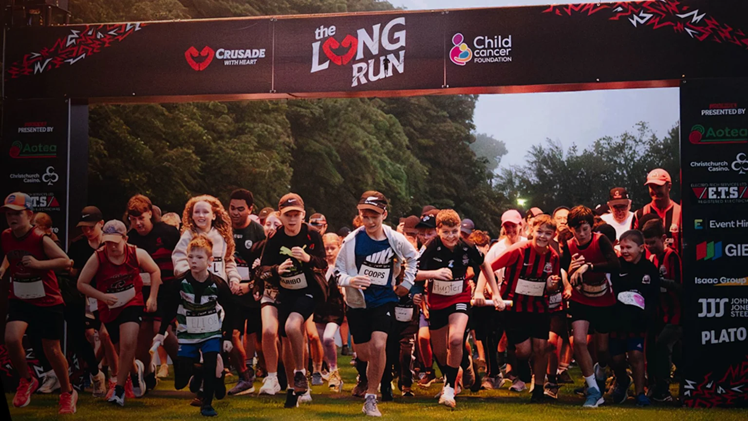Children and adults begin a race on a grassy field, running under a large banner displaying "the LONG RUN" event title and sponsor logos.
