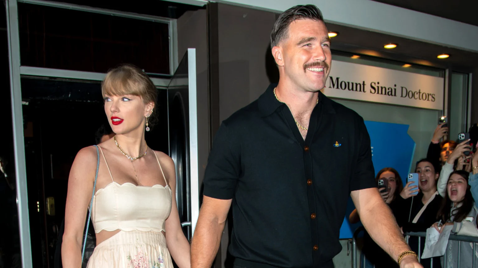 Taylor Swift in a light dress and Travis Kelce in a black shirt hold hands and smile while walking past a crowd of cheering fans.