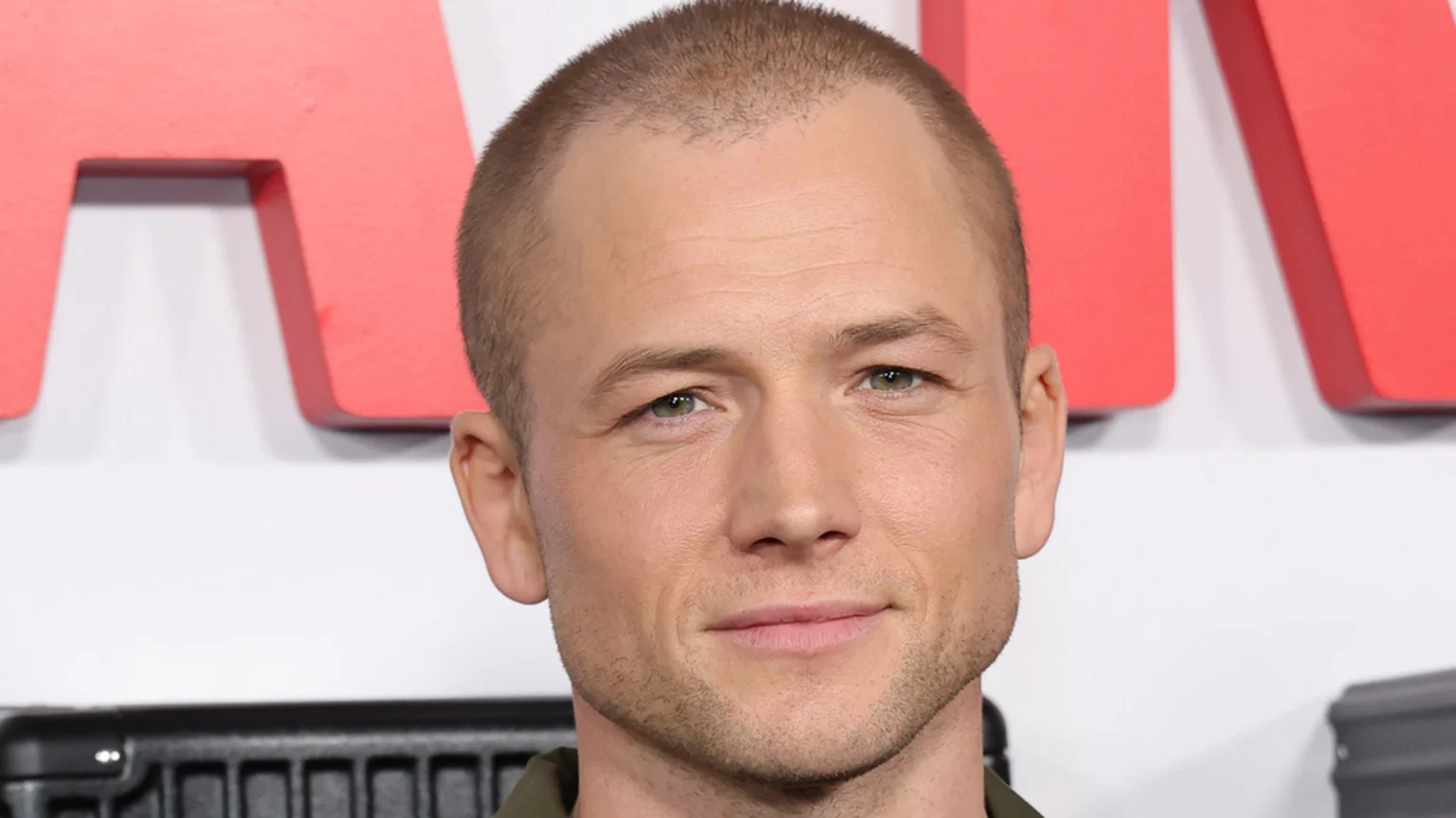 Close-up headshot of actor Taron Egerton with a buzz cut and green eyes, looking at the camera with a slight smile, with red letters partially visible in the blurred background.
