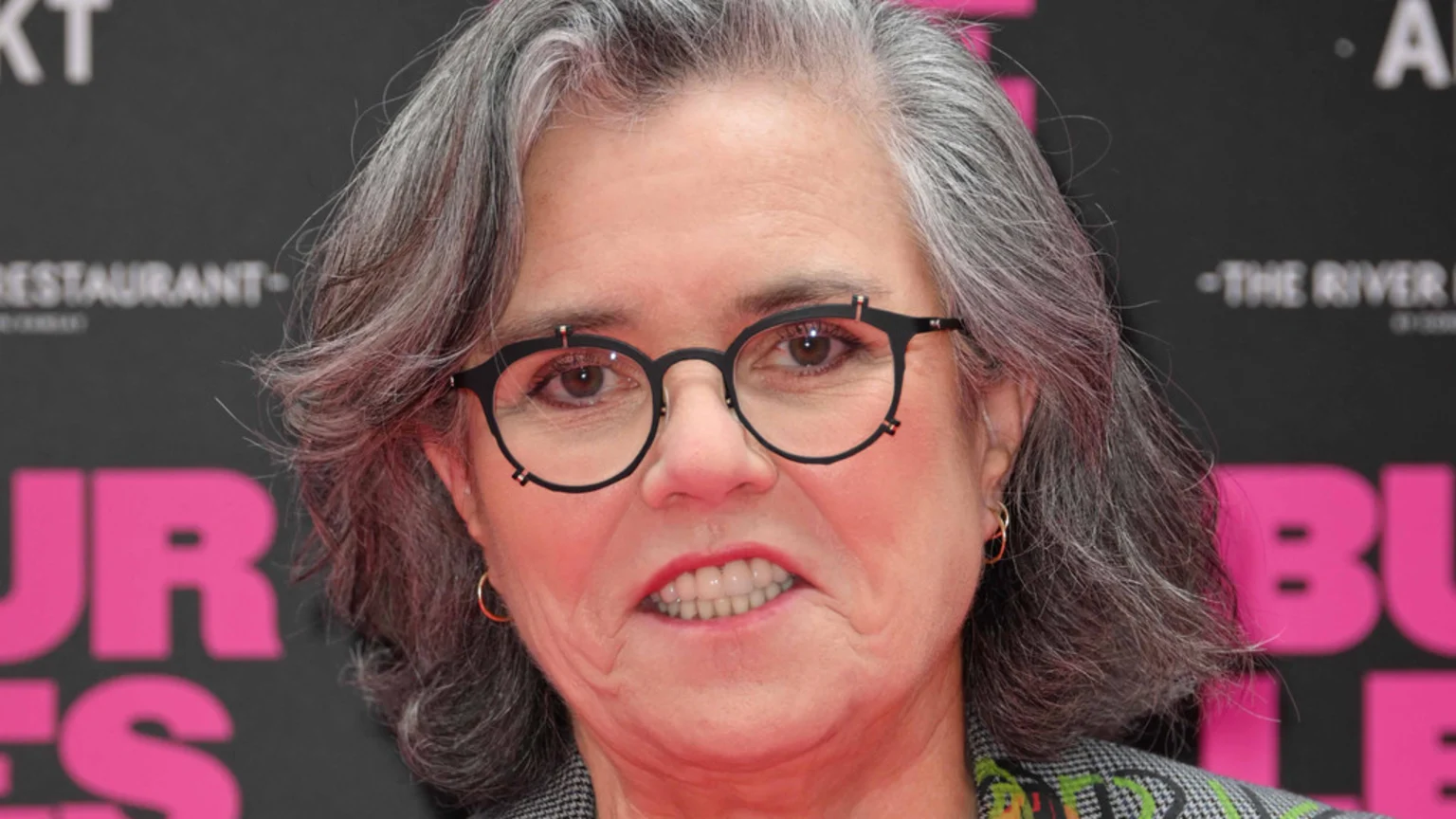 A woman with gray hair and distinct black glasses looks directly at the camera with a strained expression, revealing her teeth, against a dark background with pink text.