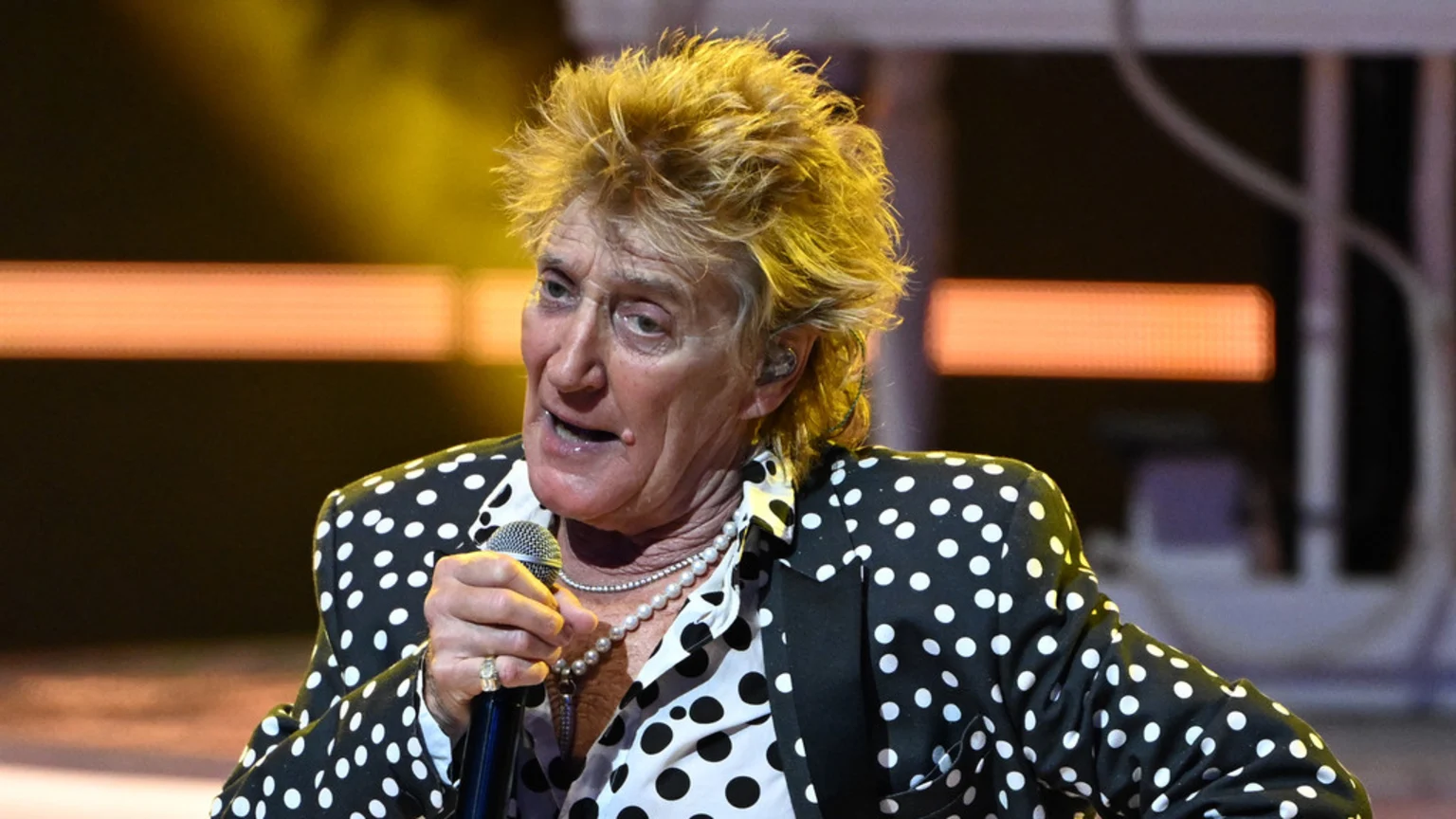Rod Stewart with spiky blonde hair, wearing a black and white polka dot suit and a pearl necklace, holds a microphone and sings on stage.