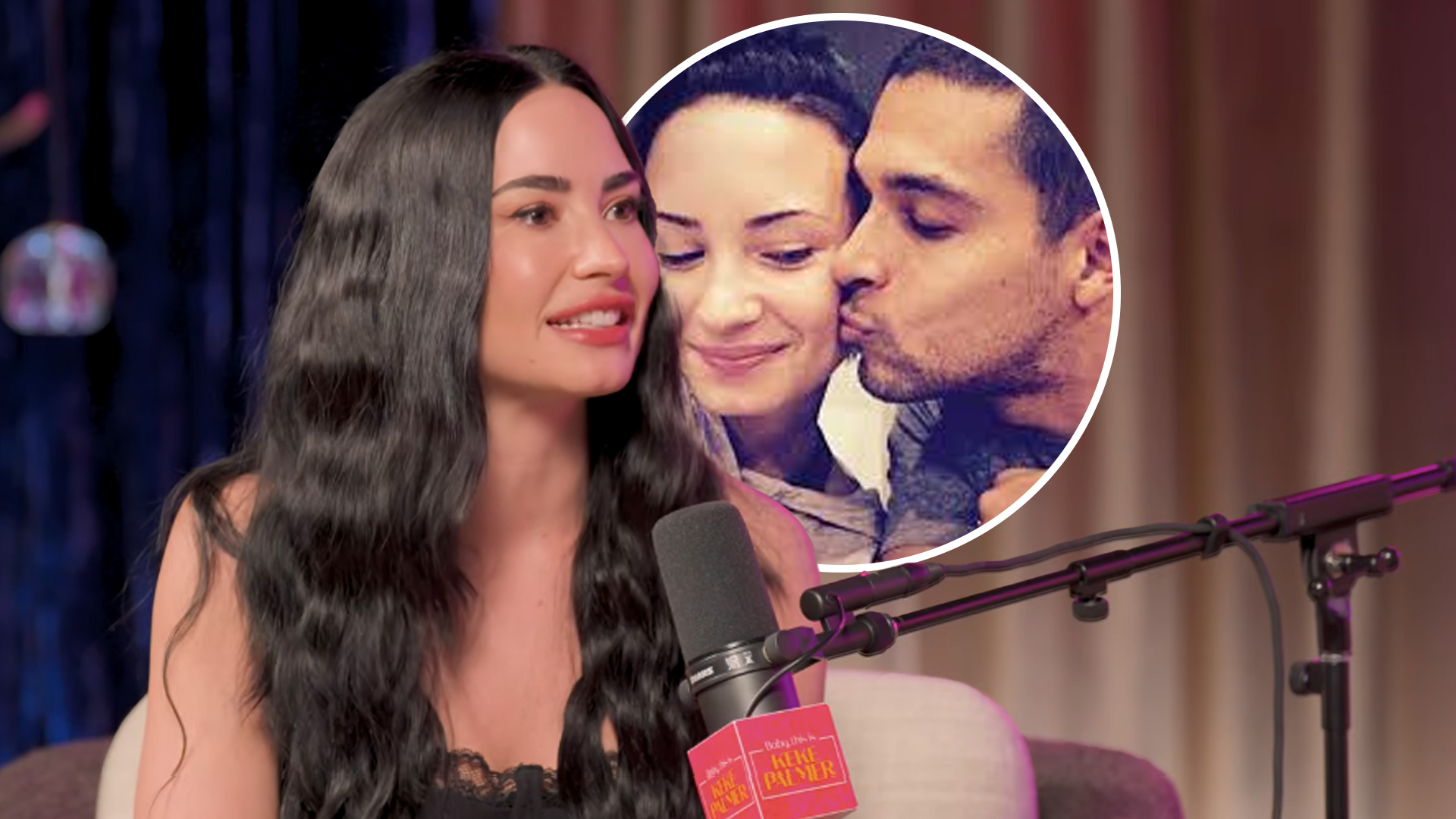 Wilmer Valderrama, in a patterned black tuxedo and bow tie, stands next to Demi Lovato, who wears a black off-the-shoulder dress and long earrings, as they pose for a photo.