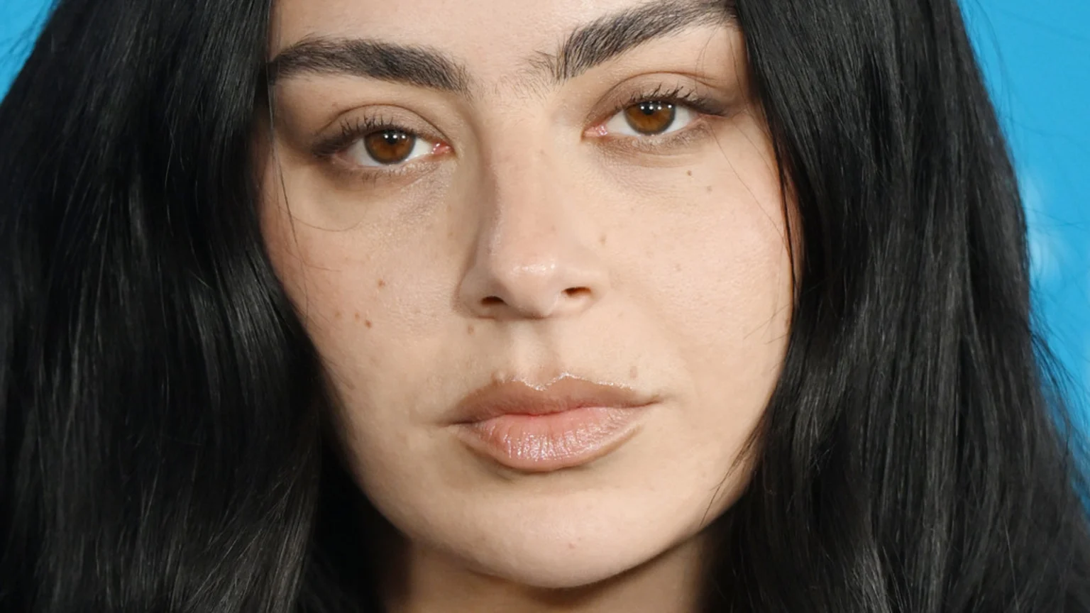 Close-up of Charli XCX's face, featuring her brown eyes, dark eyebrows, glossy lips, and dark hair, against a blue background.