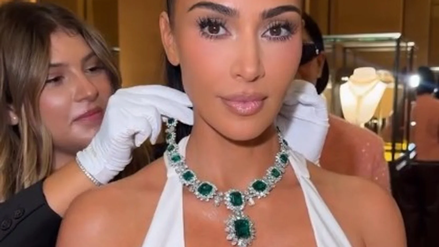 Kim Kardashian wears a large emerald and diamond necklace as a person in white gloves adjusts it around her neck, with jewelry displays in the blurred background.