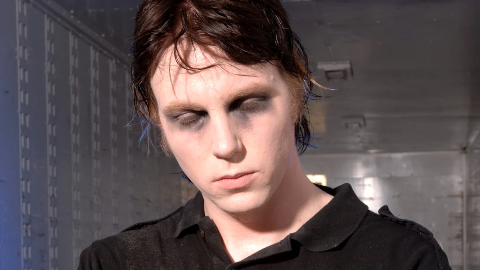 A person with pale skin, dark hair with blue streaks, and heavy black makeup around their closed eyes wears a black polo shirt, looking down in an industrial setting.