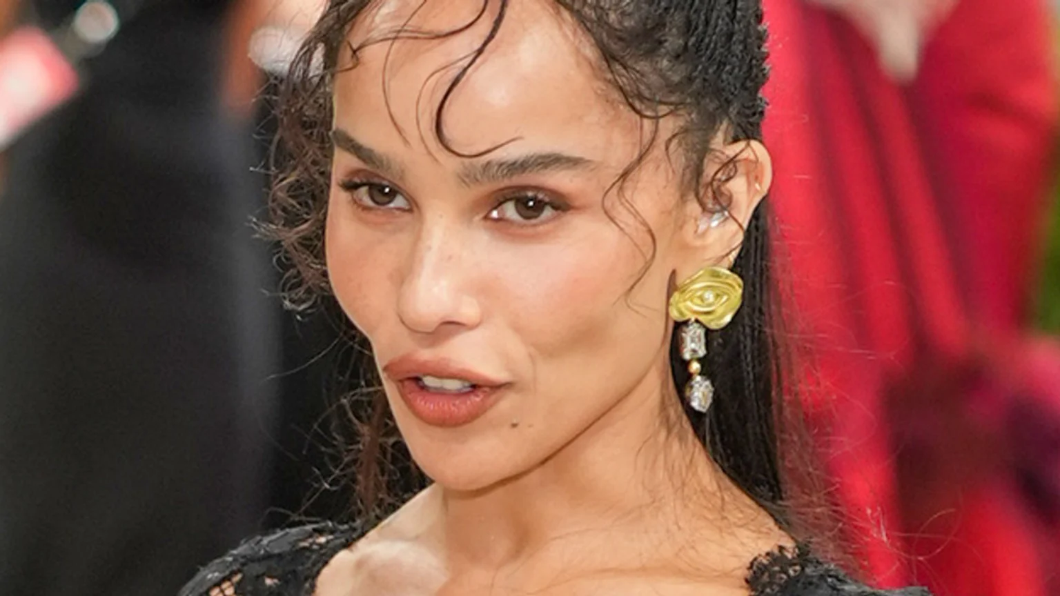 A close-up portrait of Zoe Kravitz looking directly at the camera, with dark curly hair, subtle makeup, and slightly open mouth. She wears a gold and diamond earring and a black lace top.