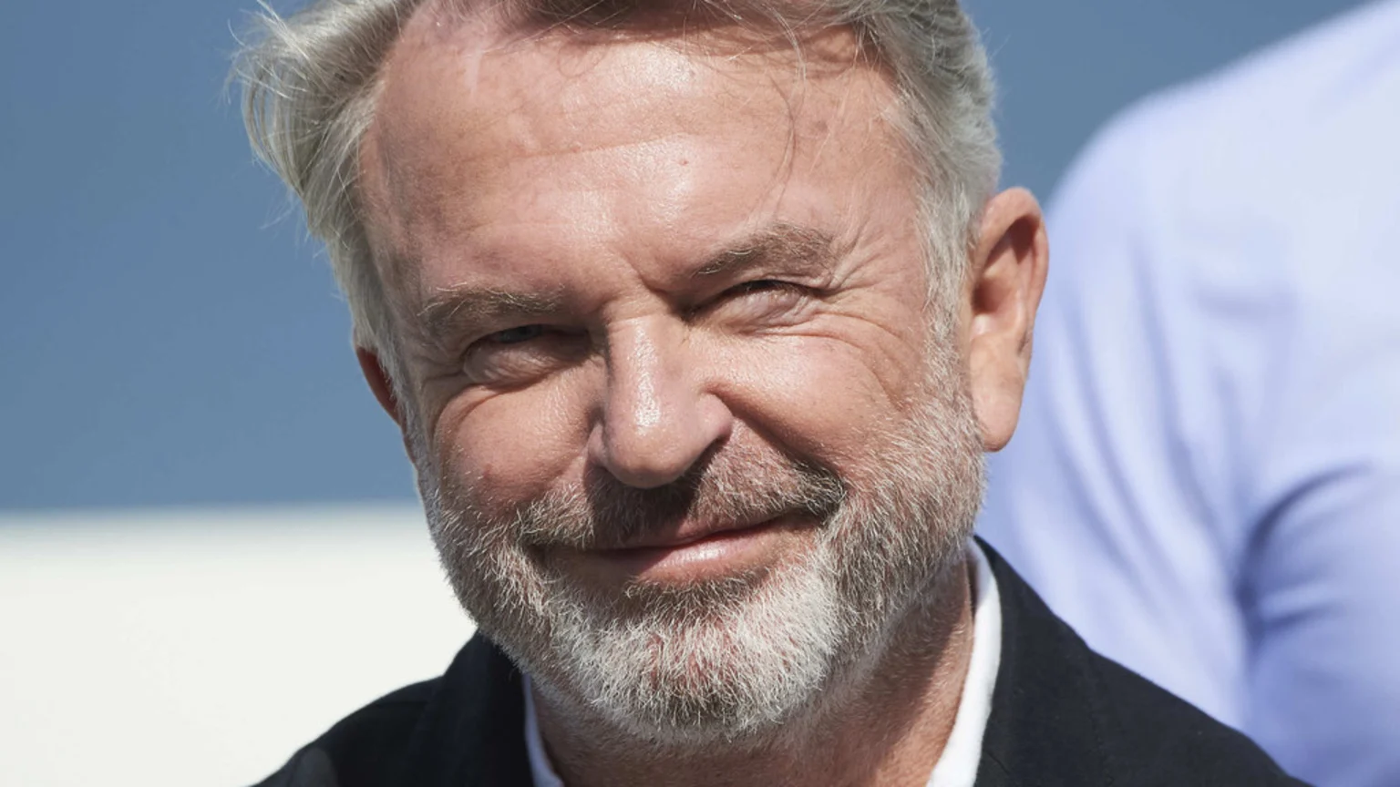 Close-up of a smiling older man with gray hair and a gray beard, wearing a dark jacket and white shirt, with a bright blue sky in the background.