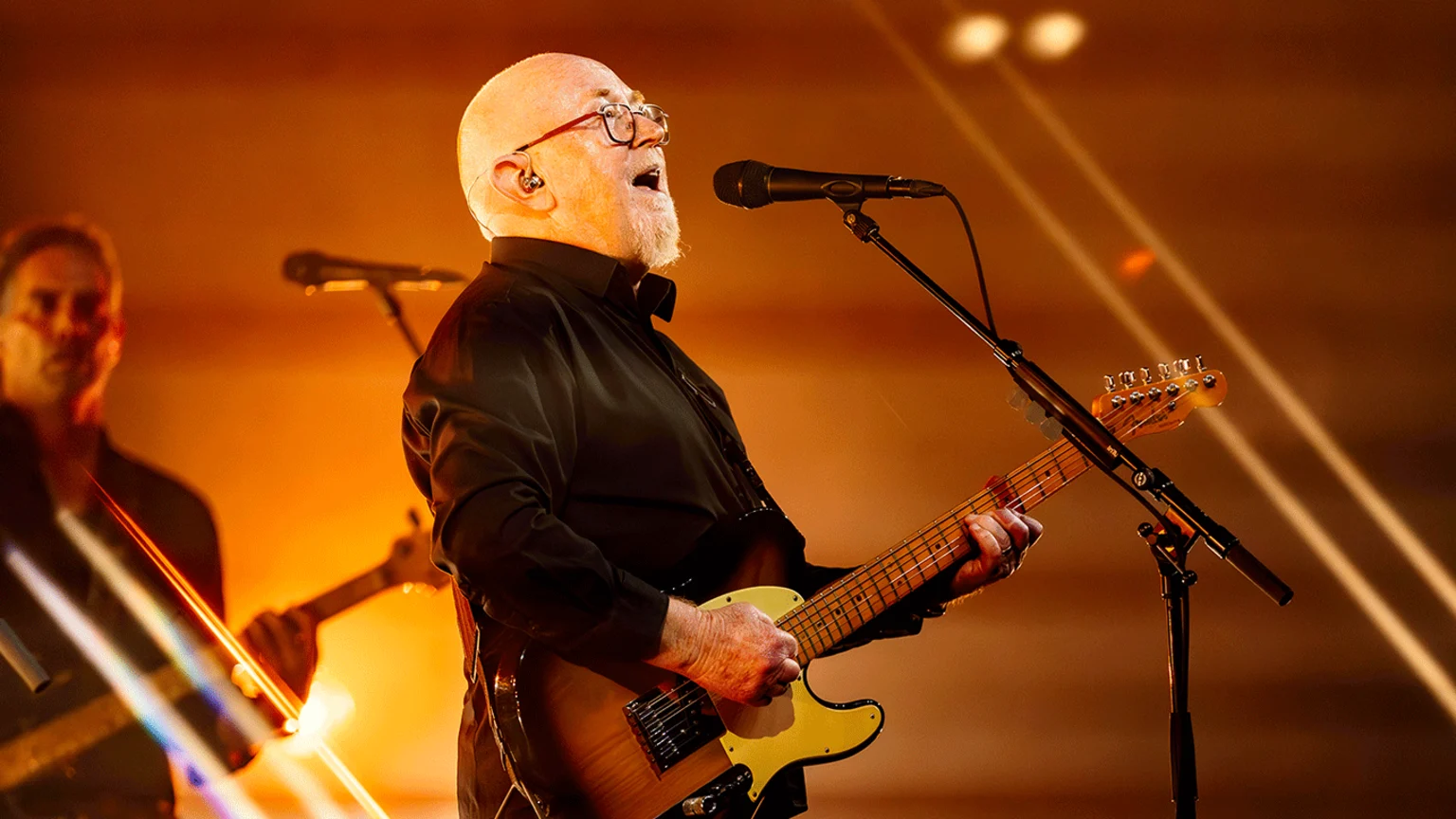 A bald, bearded man in glasses and a black shirt sings into a microphone while playing an electric guitar on a warmly lit stage, with another musician out of focus in the background.