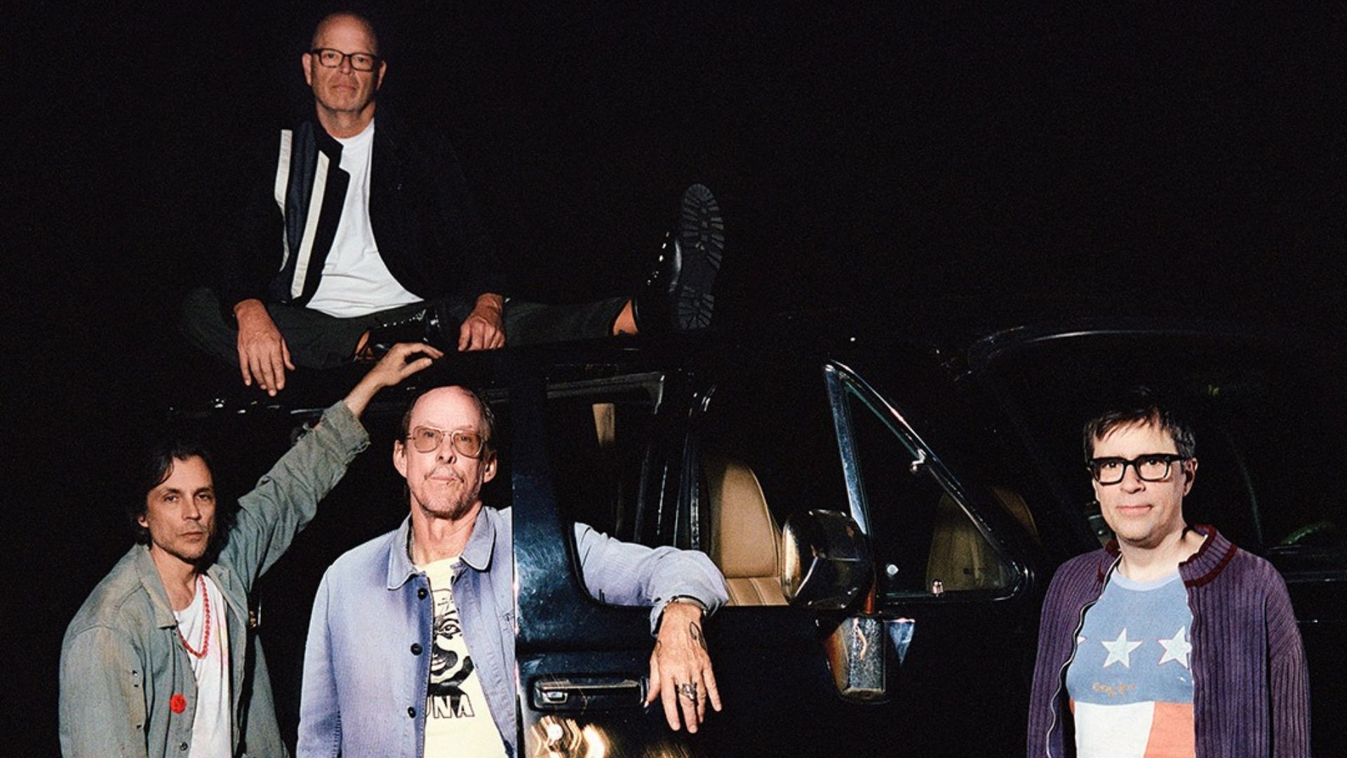 Four men, some wearing glasses, pose with a dark vehicle in a low-light setting; one sits on the roof, one leans on the side, and two stand next to it.