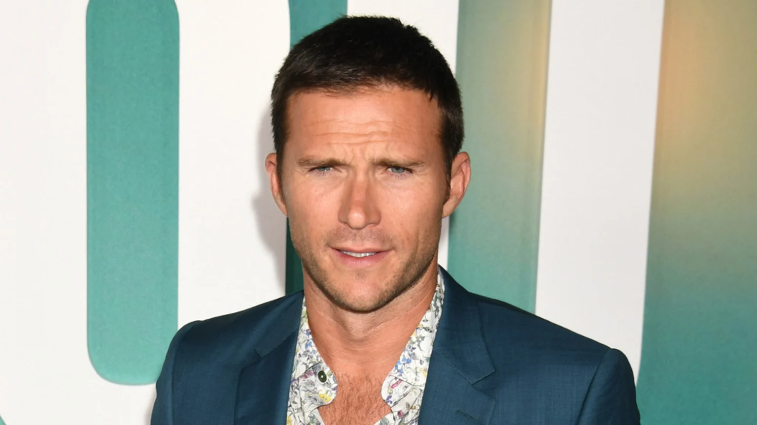 A man with short dark hair and light eyes wears a dark suit jacket over a patterned shirt, looking directly at the camera with a furrowed brow against an abstract green and white background.