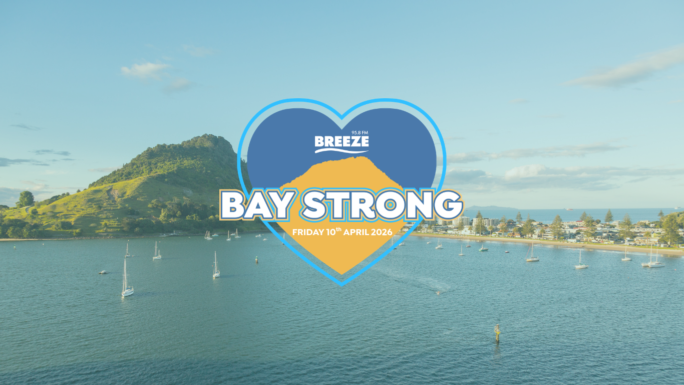 Aerial view of a bay with a green hill, a town, and many boats, overlaid with a blue and orange heart graphic that reads "95.8 FM BREEZE BAY STRONG FRIDAY 27TH MARCH 2026."
