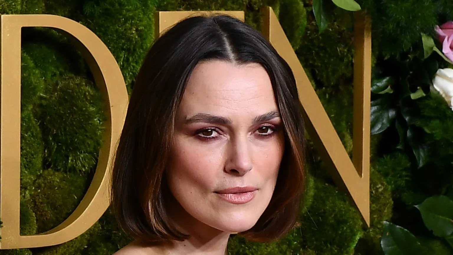 Medium shot of Keira Knightley with a bob hairstyle and center part, looking forward against a green moss wall with gold letters and flowers.