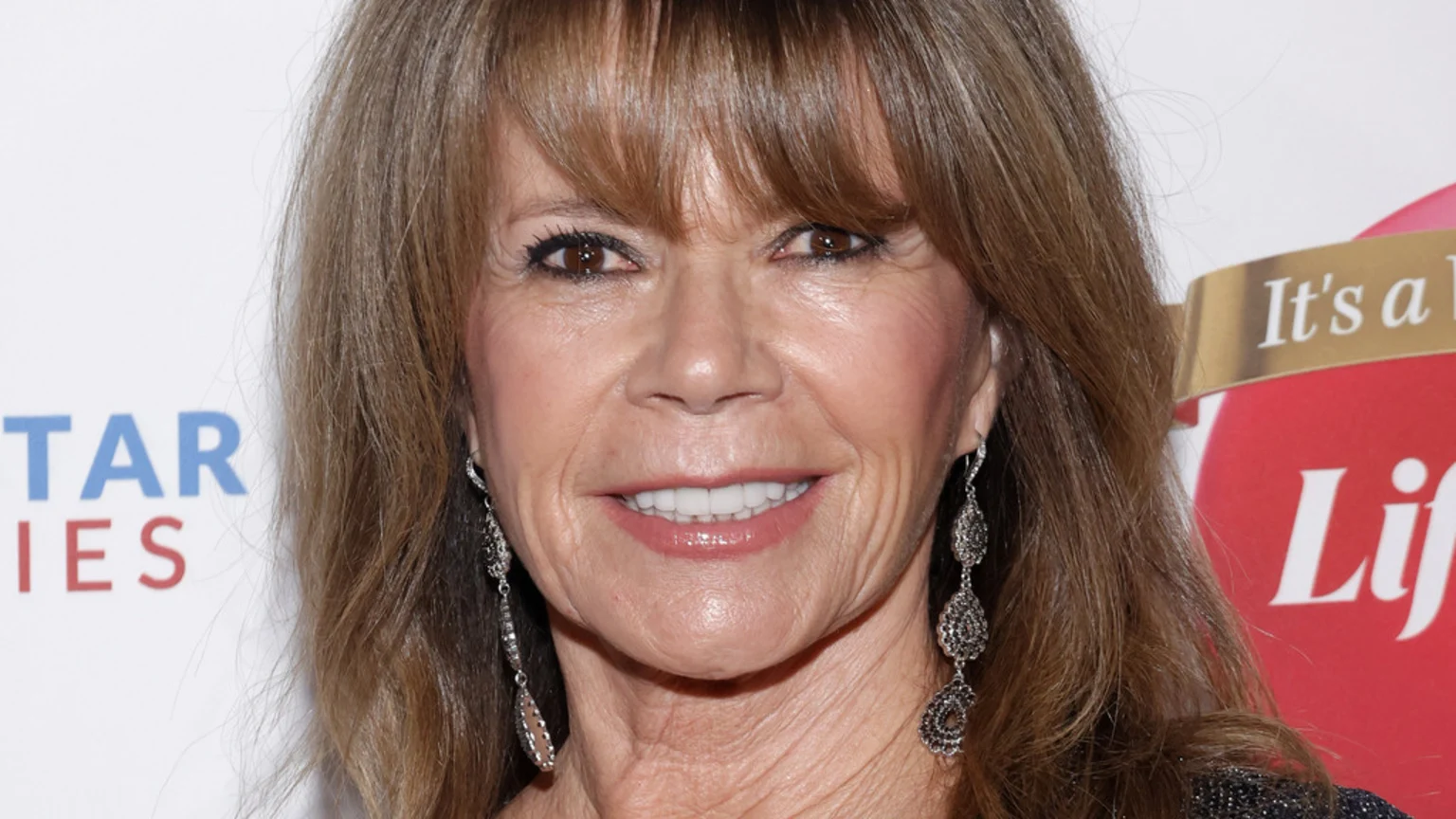 A smiling woman with light brown hair and bangs, wearing long ornate dangling earrings, looks forward, with blurred event logos partially visible in the background.
