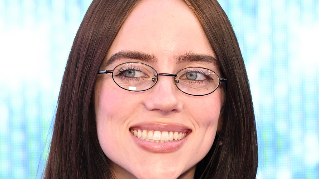 A close-up portrait of Billie Eilish smiling broadly, wearing small oval glasses with her light blue eyes looking up and to the sides, framed by long dark hair against a blurred blue background.