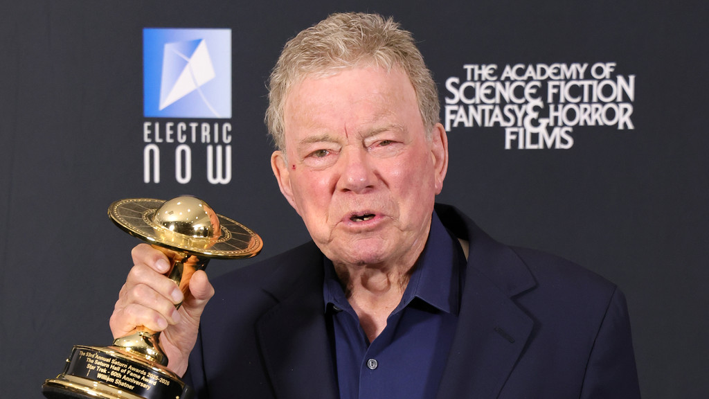 Elderly William Shatner holds his golden Saturn Hall of Fame Award, shaped like a UFO, against a black background with logos for Electric Now and The Academy of Science Fiction, Fantasy & Horror Films.