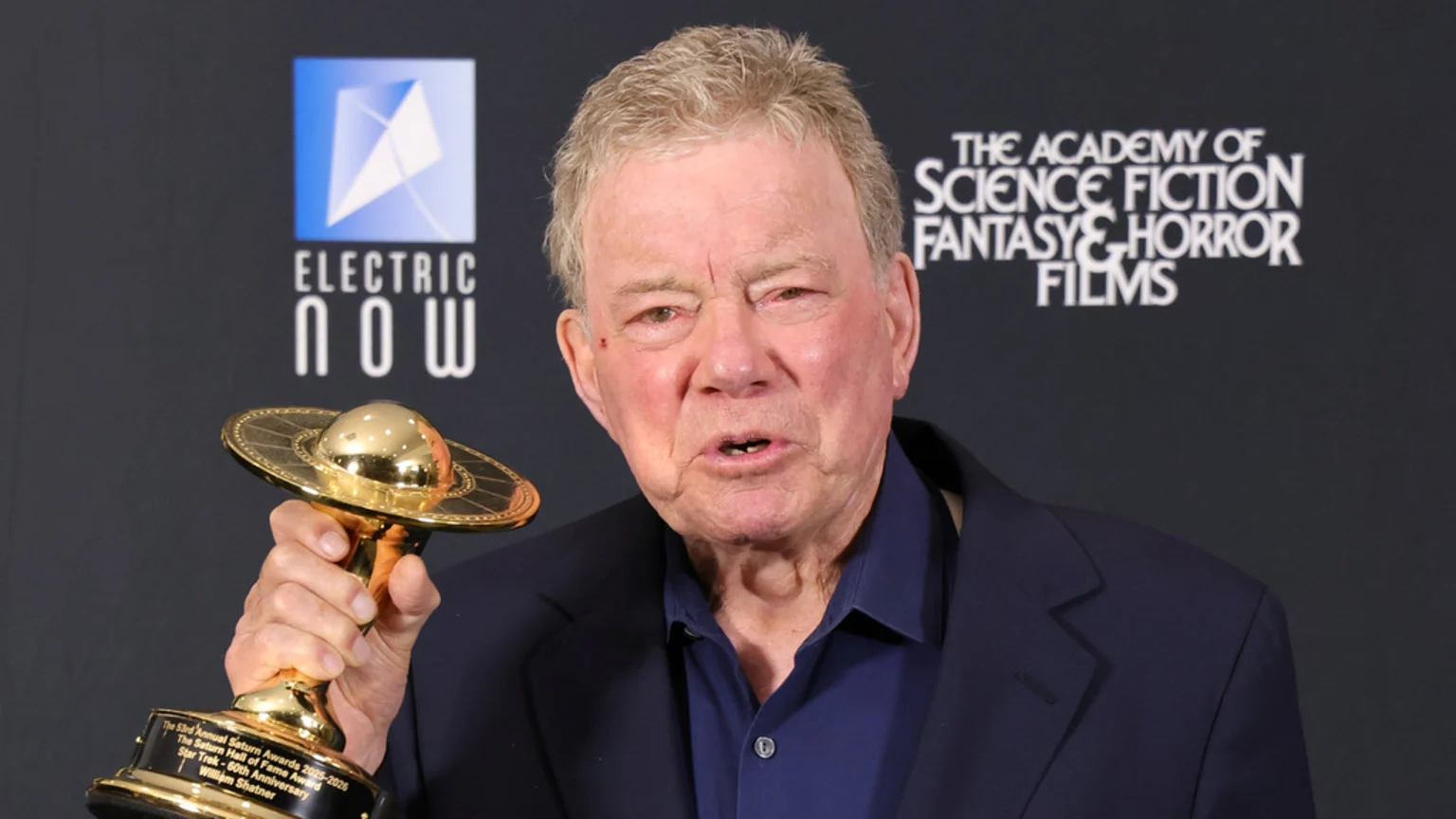 Elderly William Shatner holds his golden Saturn Hall of Fame Award, shaped like a UFO, against a black background with logos for Electric Now and The Academy of Science Fiction, Fantasy & Horror Films.
