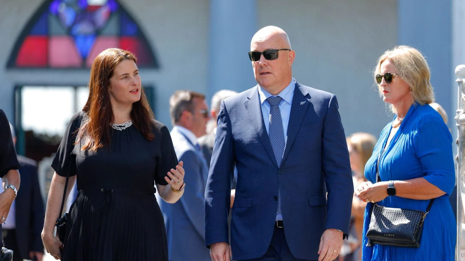 A bald man in a blue suit and sunglasses walks between two women, one in a black dress with a pearl necklace, and another in a blue dress, outdoors with a building in the background.
