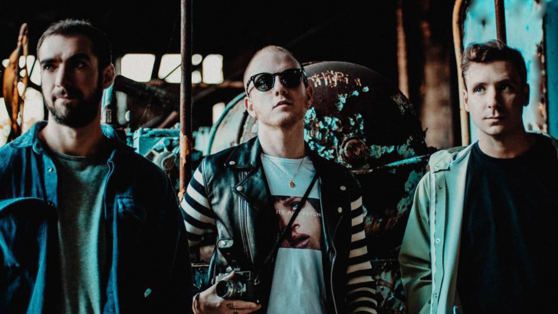 Three men stand in an industrial-looking space, with the central figure wearing sunglasses and a leather jacket while holding a camera, flanked by two others.