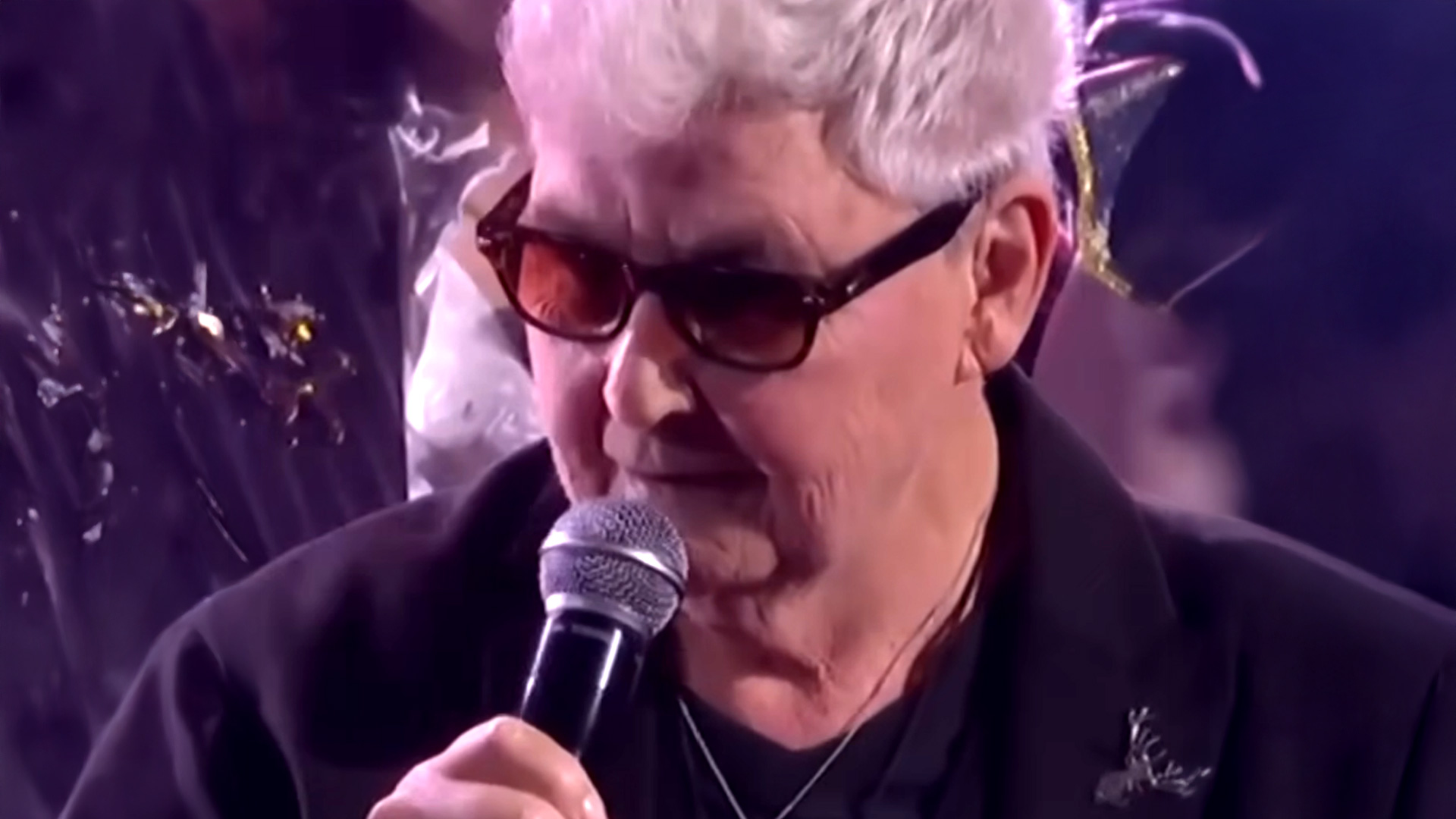 An older person with white hair and sunglasses, wearing a dark jacket with a deer-shaped brooch, speaks into a microphone.
